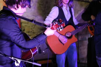 Photo d'un concert avec au centre de l'attention, une personne blanche qui joue de la guitare sèche, en chemise blanche et veston noire, et au premier plan, une autre personne blanche jouant de la guitare sèche, en veste noire.